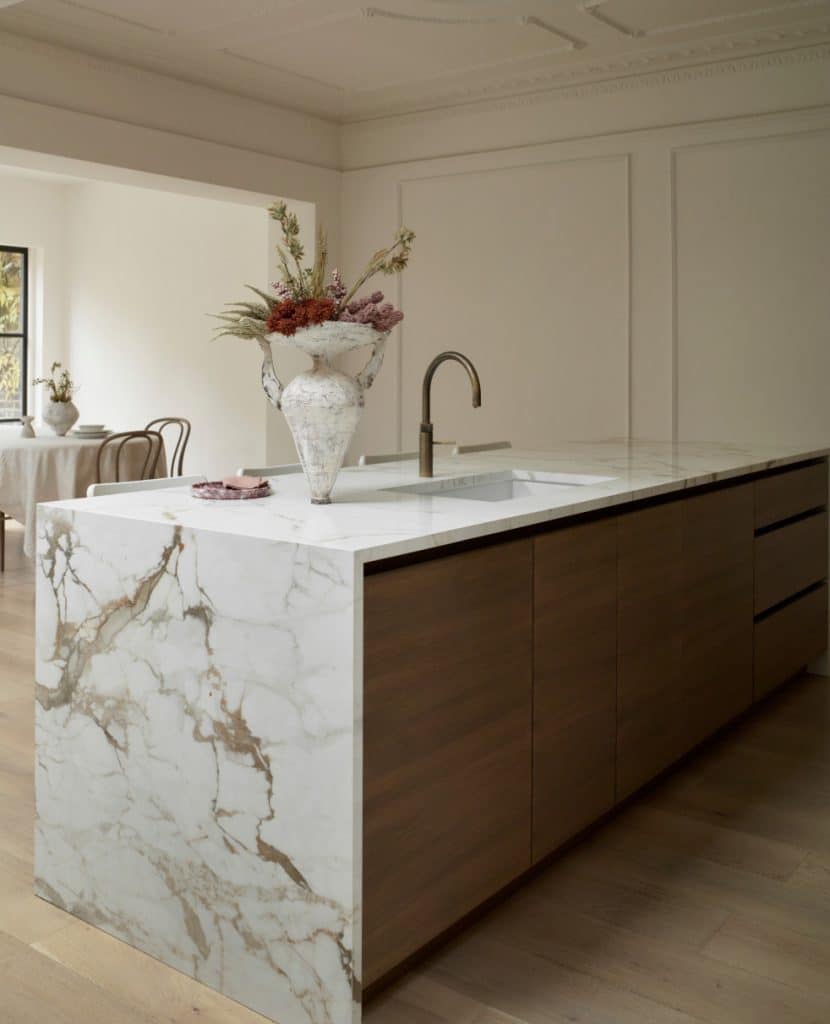 A modern kitchen island featuring a white marble countertop with prominent grey and gold veining and a waterfall edge. The island includes a built-in sink with a sleek, curved brass faucet, dark wood lower cabinetry, and a large decorative vase filled with dried flowers sitting on top.